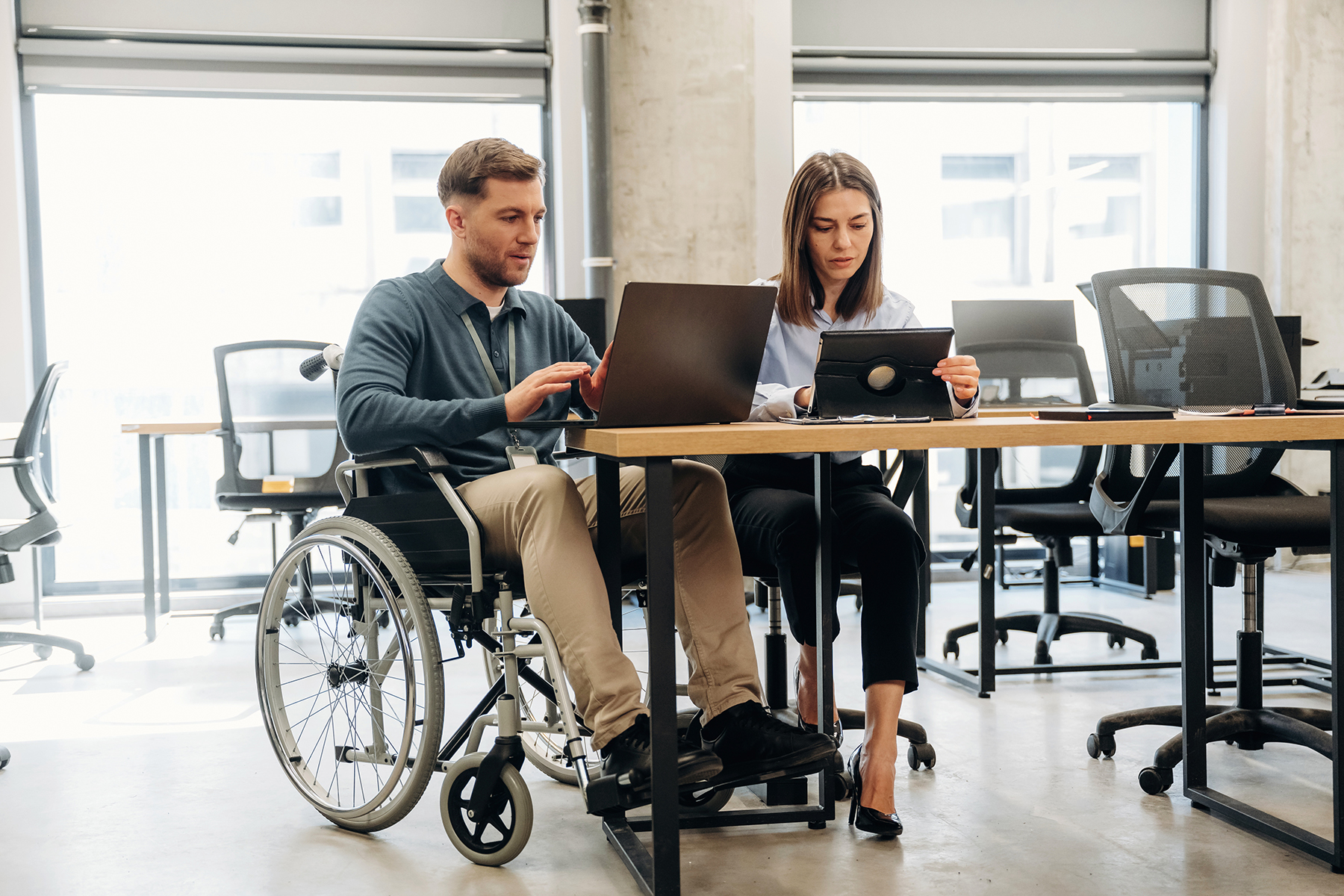 Male in wheelchair and female colleague work together using a laptop and tablet in a contemporary office setting with empty chairs and large windows